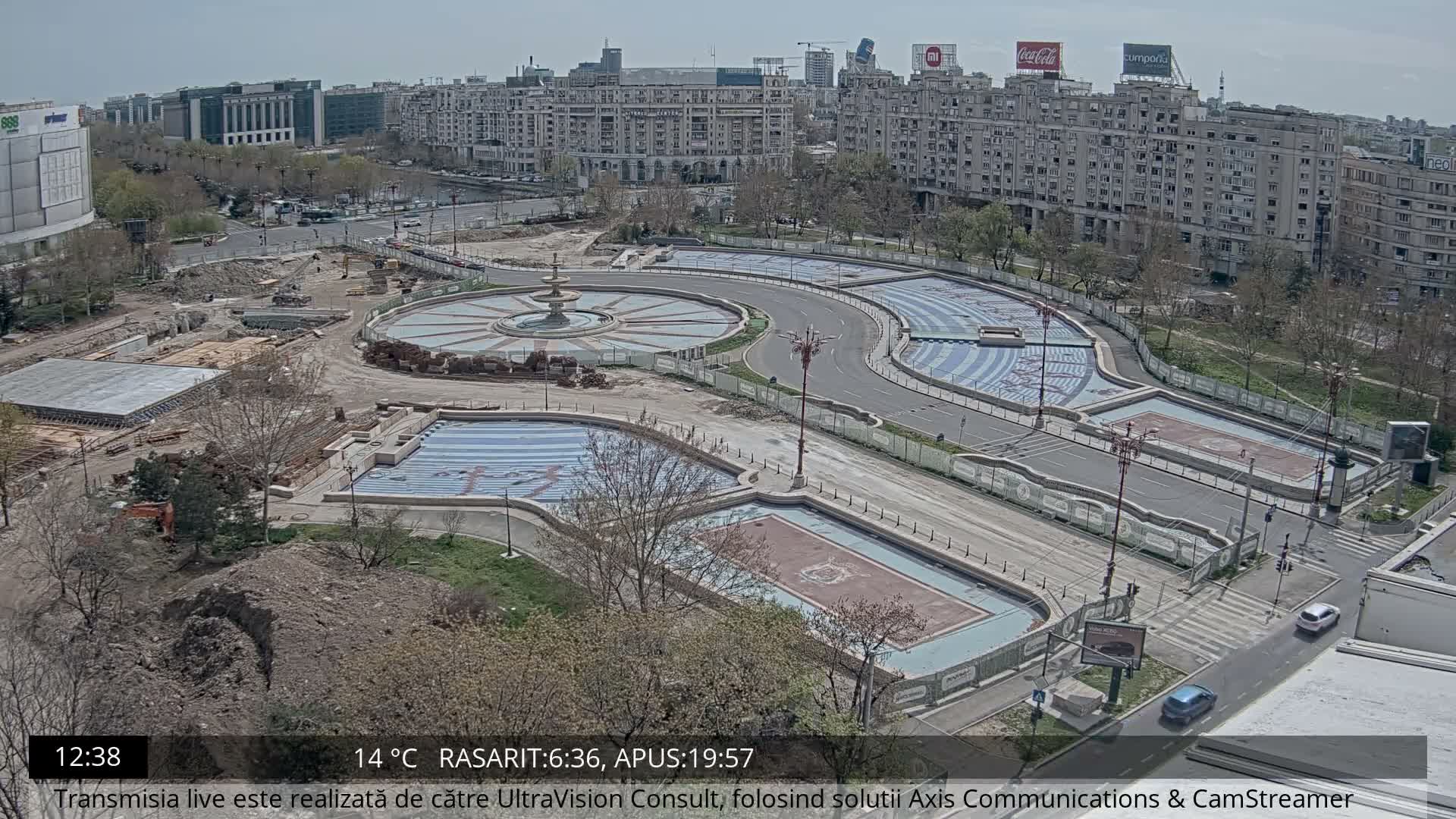 An aerial view captures a bustling city intersection undergoing construction, featuring multiple roads with moving traffic, several ornate but seemingly empty fountain pools, and numerous buildings under an overcast and damp sky.