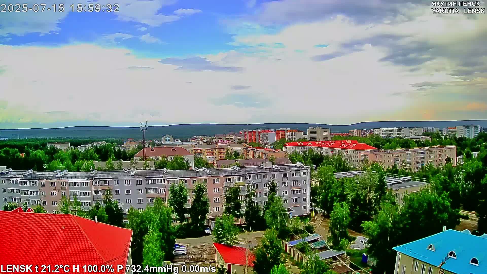 A partly cloudy day reveals a city of multi-story apartment buildings surrounded by green trees and a distant mountain range.