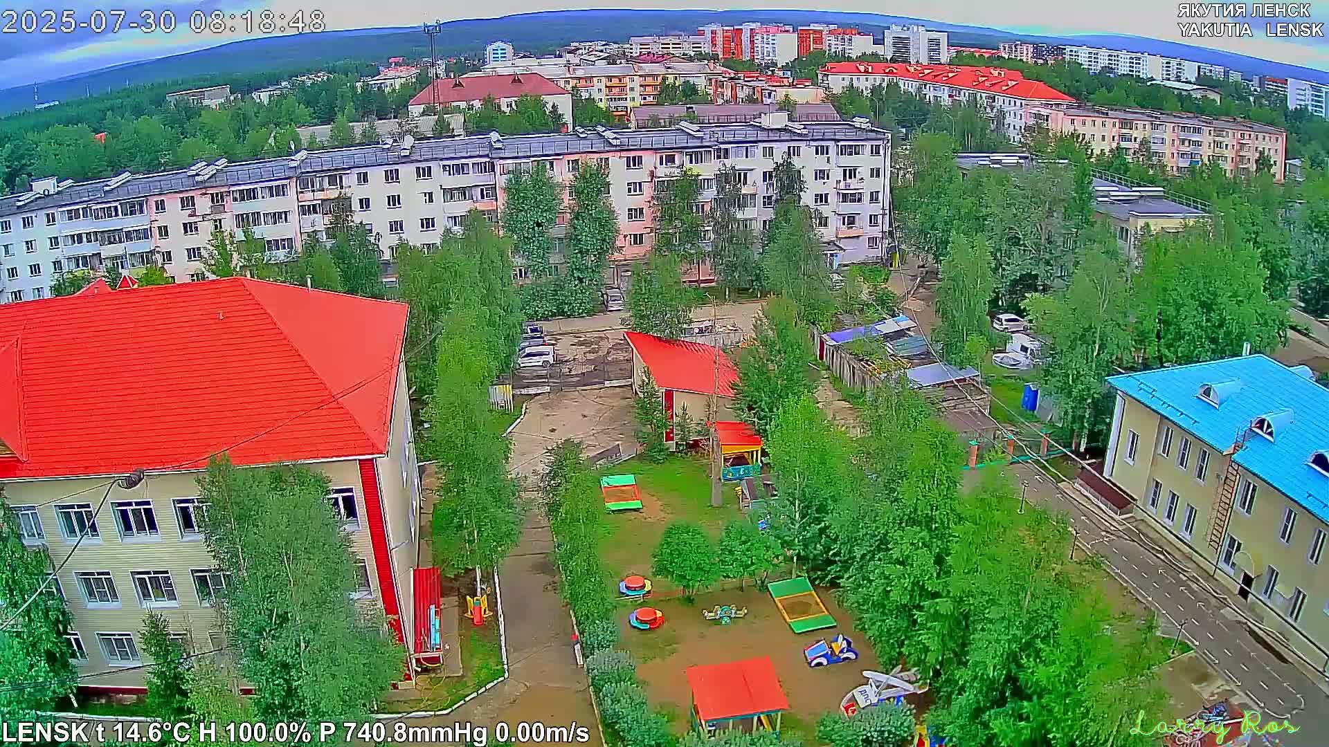 A partly cloudy day reveals a city of multi-story apartment buildings surrounded by green trees and a distant mountain range.