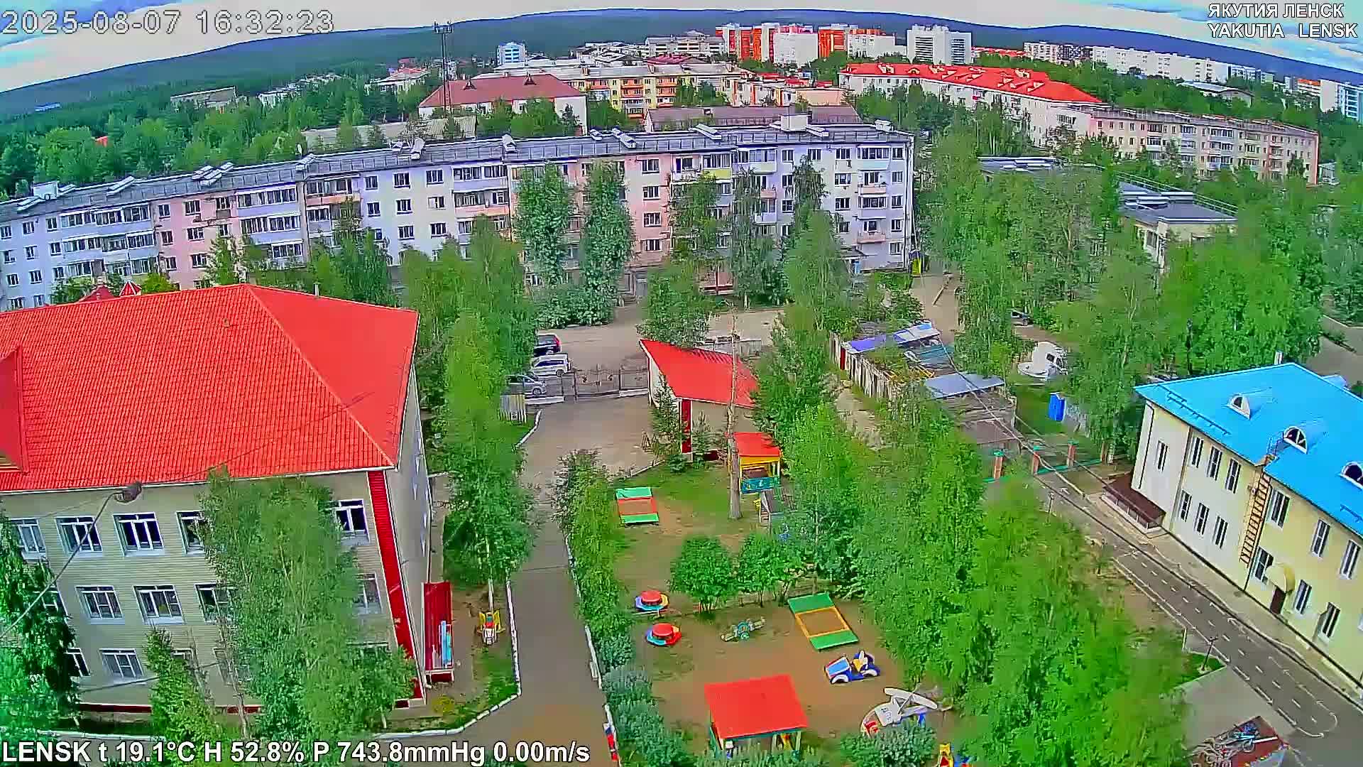 An aerial view shows a residential area with apartment buildings and a playground, under partly cloudy skies.