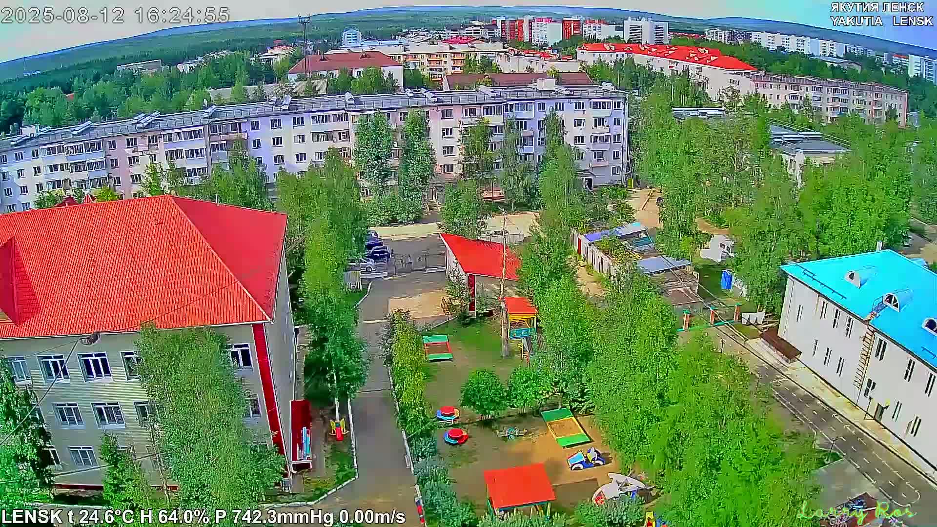 An aerial view shows a residential area with apartment buildings, a playground, and lush green trees on a partly cloudy day.