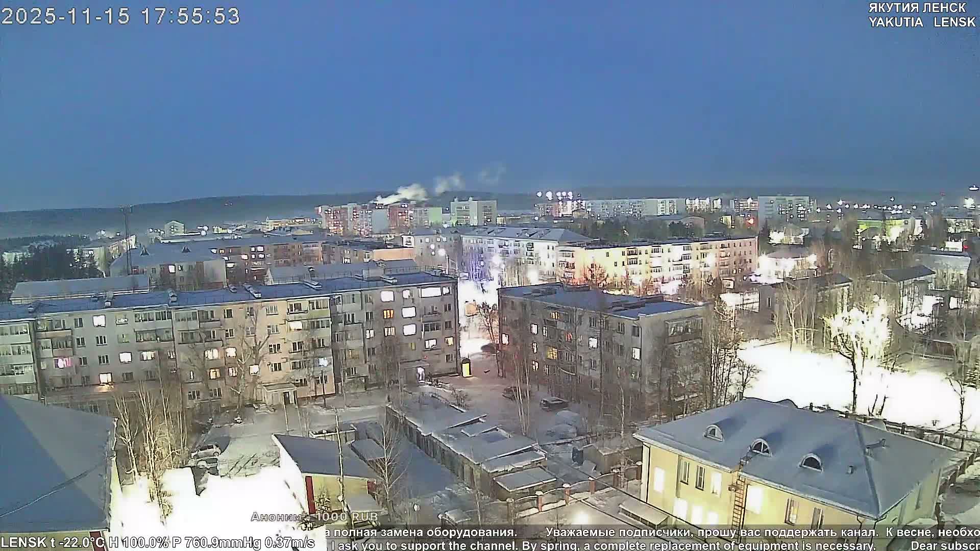 The image displays a snow-covered city at dusk or early evening, with street and building lights glowing under a clear, dark blue sky, indicating cold winter weather.