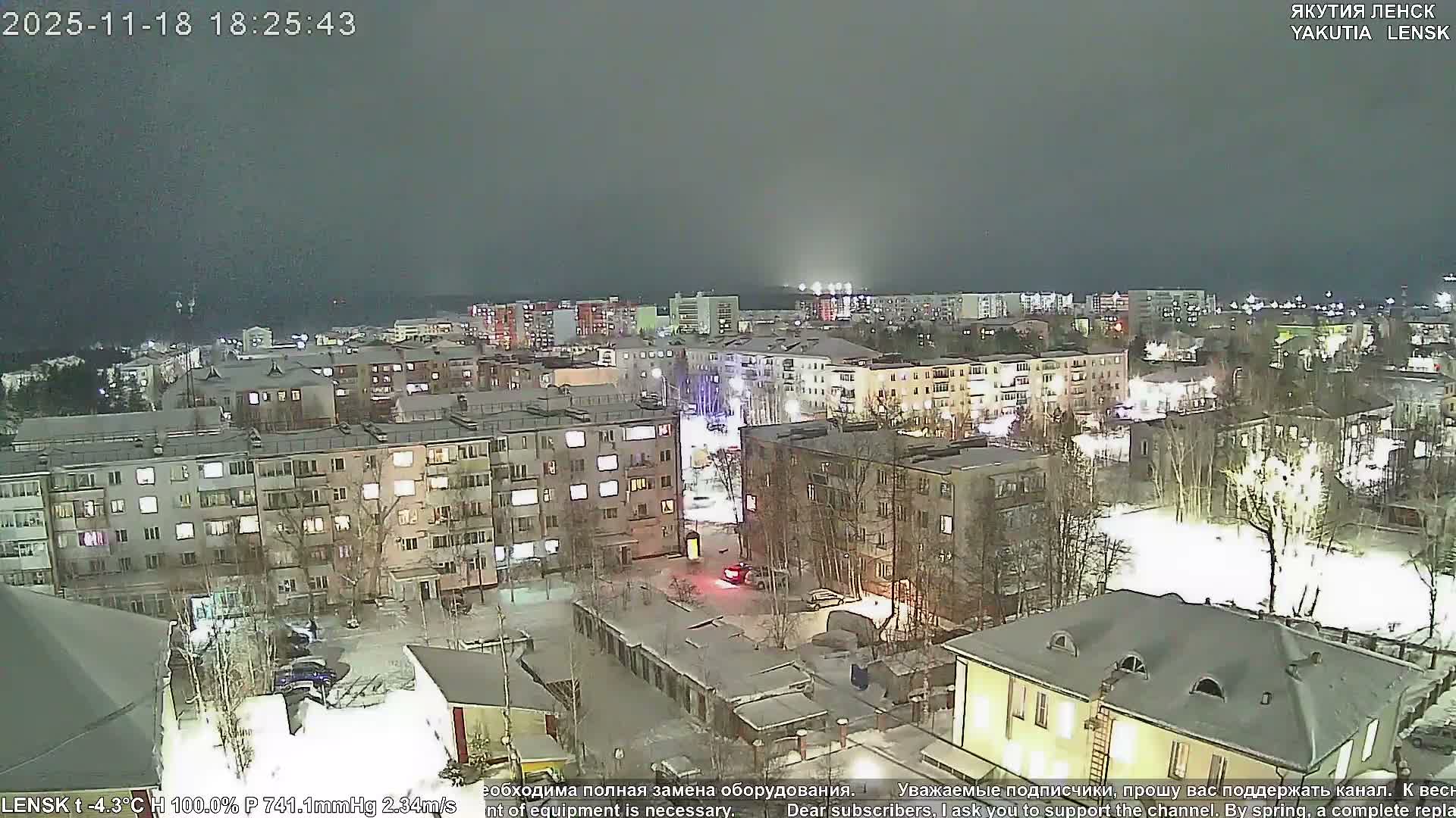 The image displays a snow-covered city at dusk or early evening, with street and building lights glowing under a clear, dark blue sky, indicating cold winter weather.