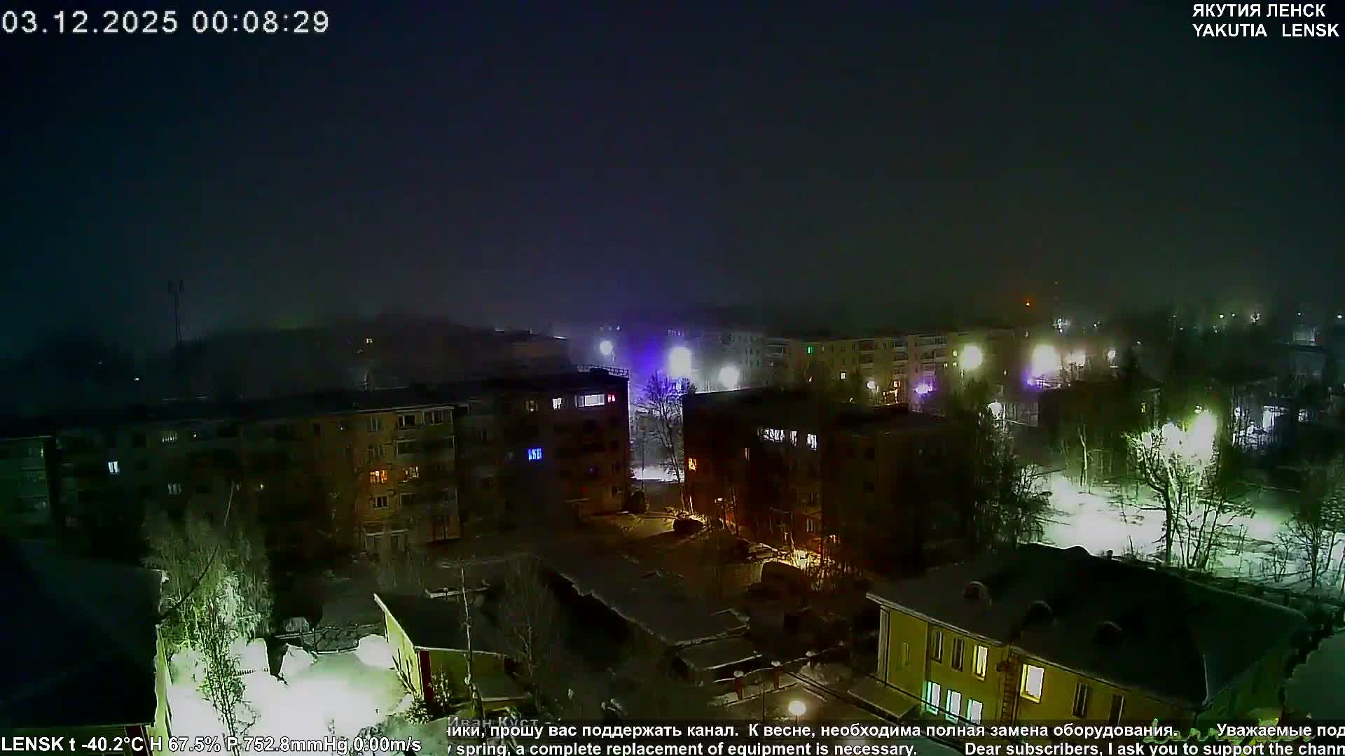 A dark, hazy night scene overlooks a snow-covered residential area with buildings and bare trees, their forms softened by diffused artificial lights, hinting at very cold and possibly foggy weather.