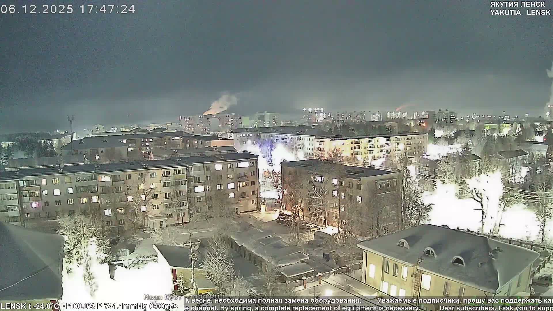 An aerial view captures a snow-covered city at night, with illuminated apartment buildings, bare trees, and distant industrial plumes, all under a dark, hazy sky in frigid winter conditions.