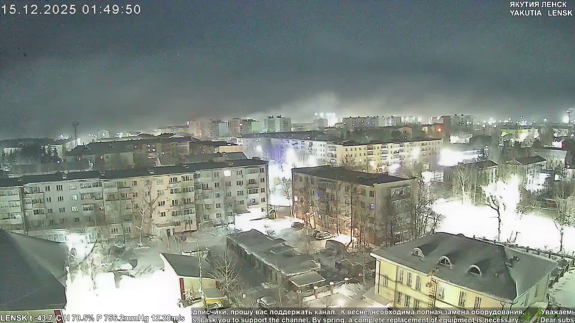 An aerial view captures a snow-covered city at night, with illuminated apartment buildings, bare trees, and distant industrial plumes, all under a dark, hazy sky in frigid winter conditions.