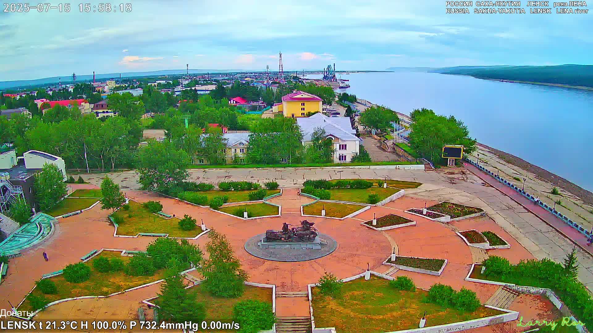 An elevated view of a town square with a statue in the center, next to a large river under a mostly cloudy sky.