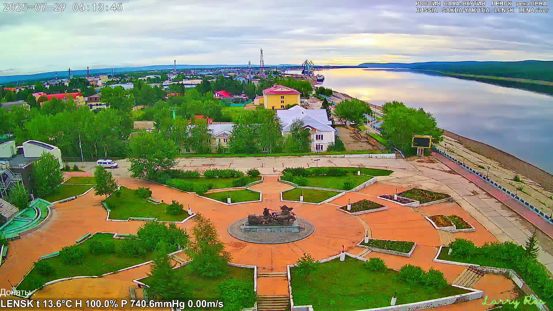 An aerial view of a town square with a statue at its center, next to a river under mostly cloudy skies.