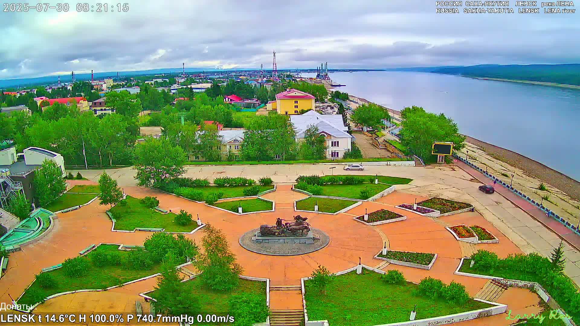 An aerial view of a town square with a statue at its center, next to a river under mostly cloudy skies.