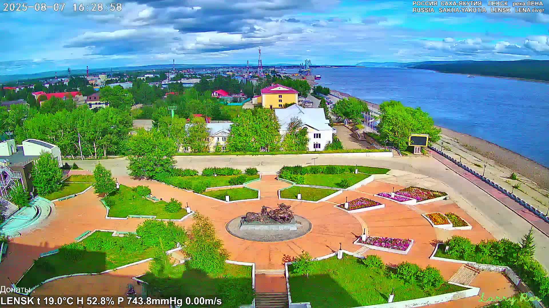 An elevated view shows a town square with a central monument, surrounded by flowerbeds and green spaces, next to a wide river under partly cloudy skies.