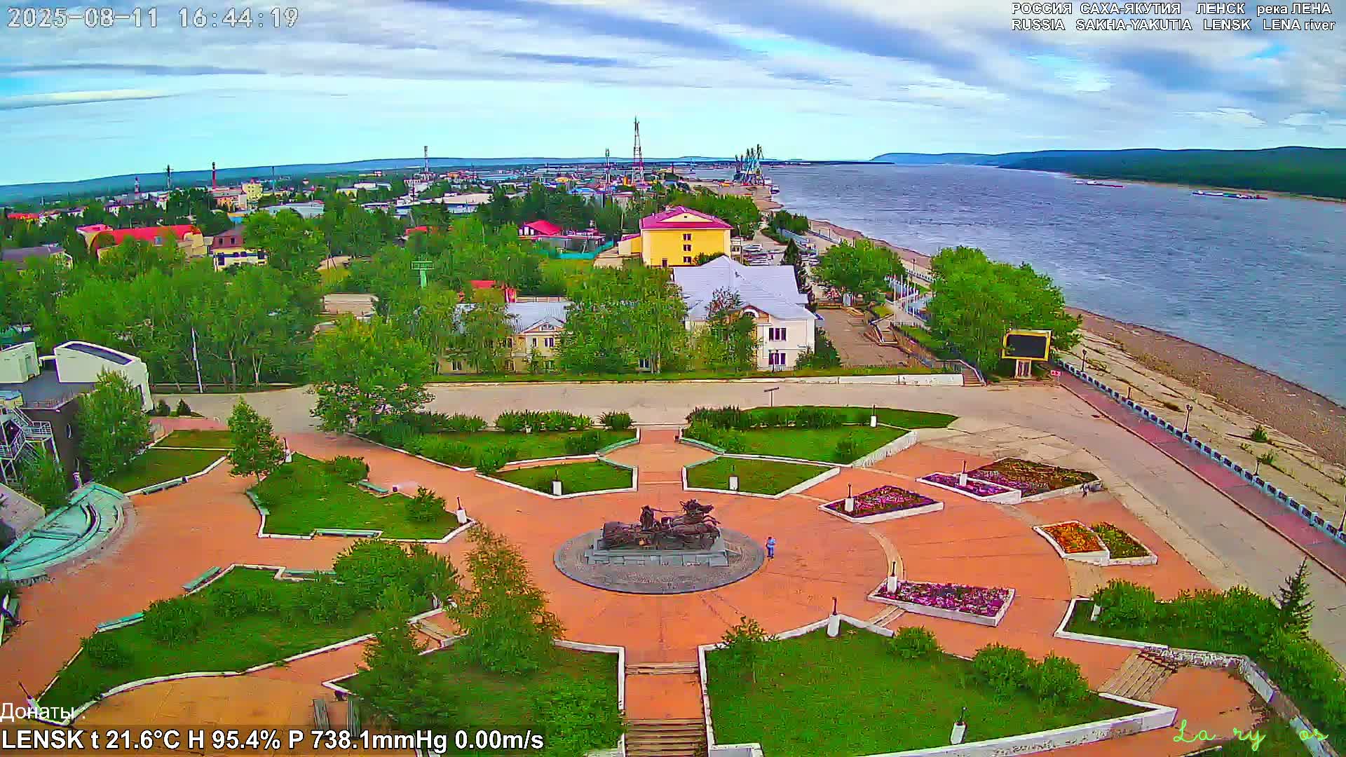 An elevated view shows a town with a park featuring a monument in its center, situated next to a wide river under partly cloudy skies.
