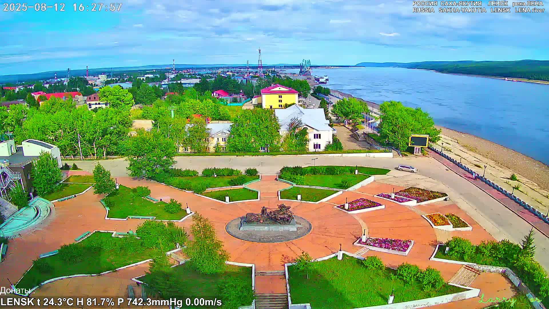 An aerial view shows a town with a park featuring a circular monument, situated near a wide river under partly cloudy skies.