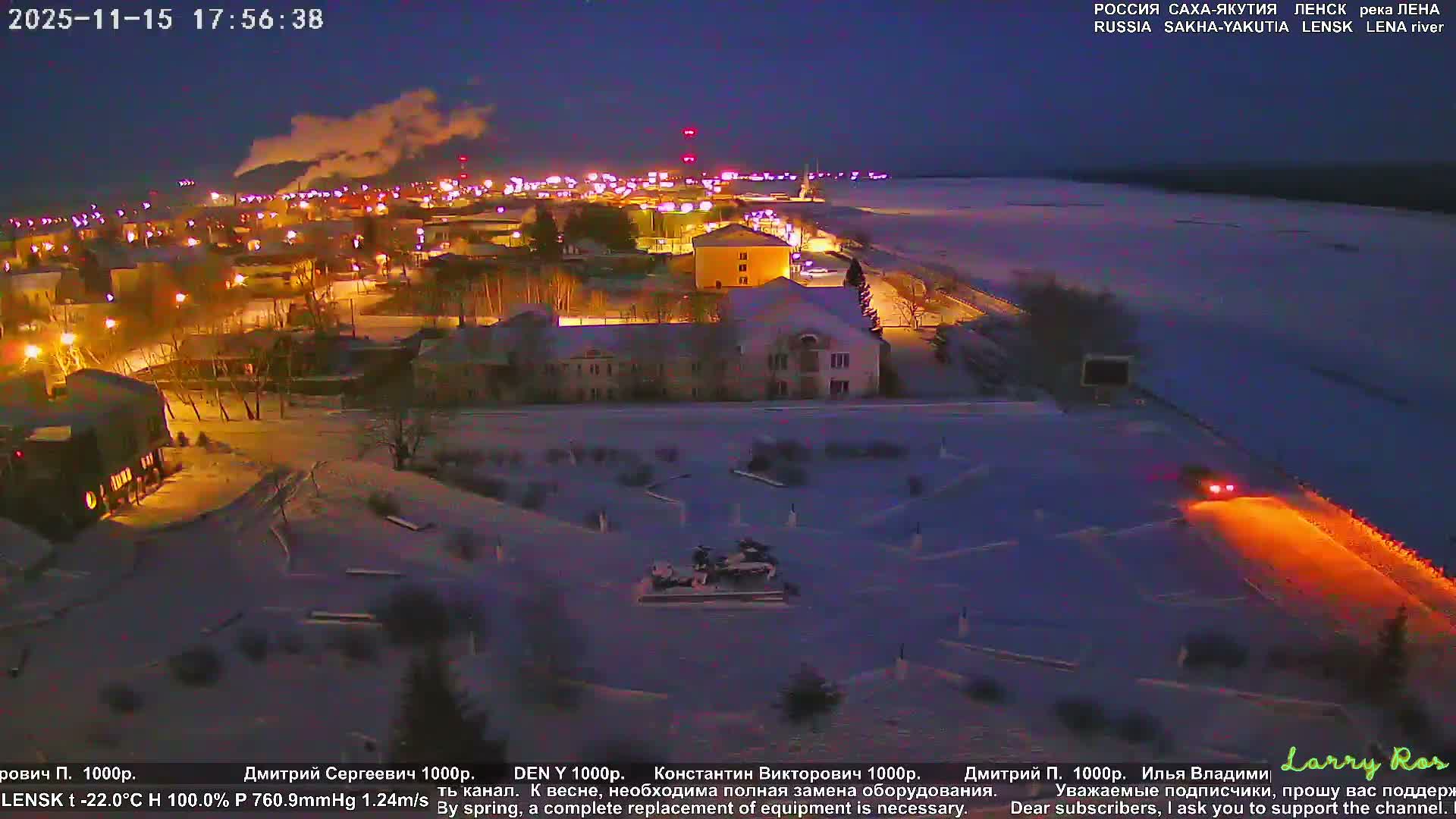 A snow-covered town on the bank of a frozen river is brightly lit on a very cold, clear winter night, with steam rising from distant industrial structures and vehicles moving on illuminated roads.