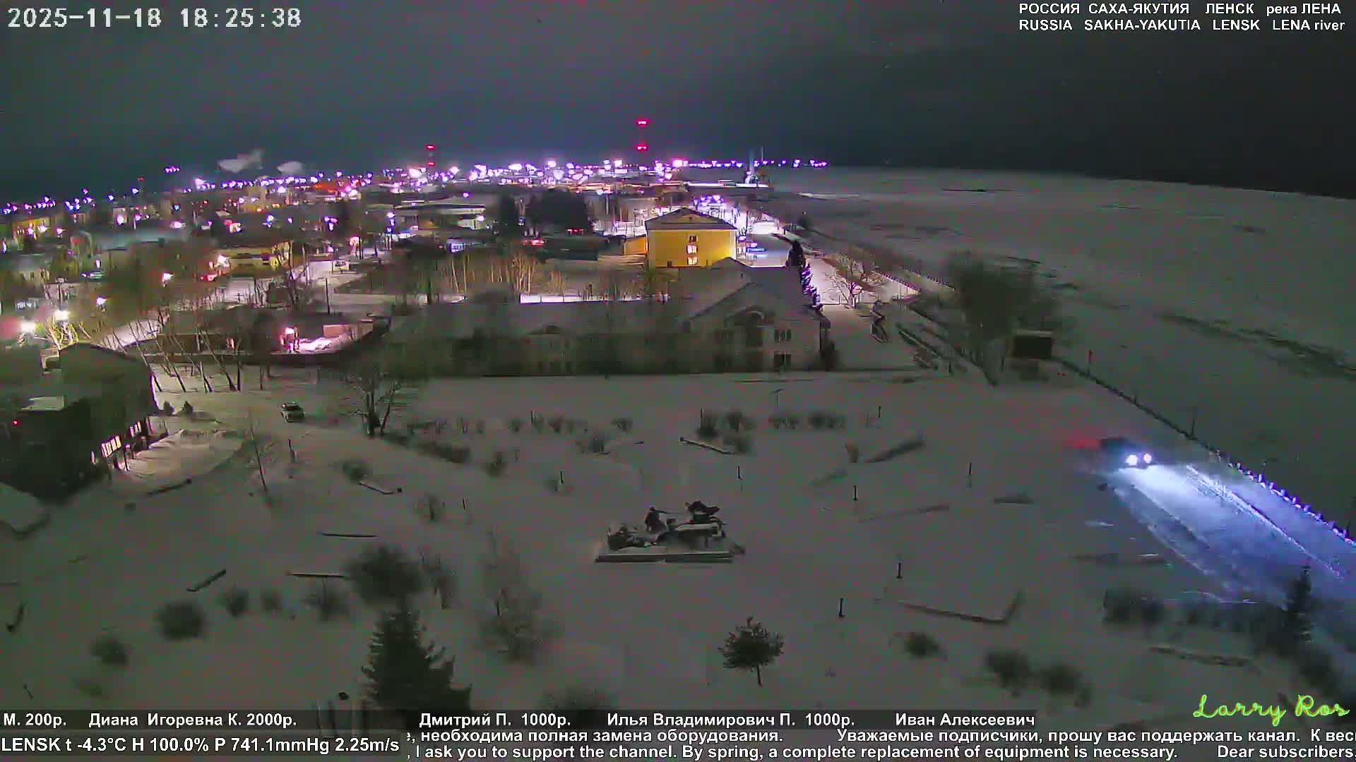 A snow-covered town on the bank of a frozen river is brightly lit on a very cold, clear winter night, with steam rising from distant industrial structures and vehicles moving on illuminated roads.