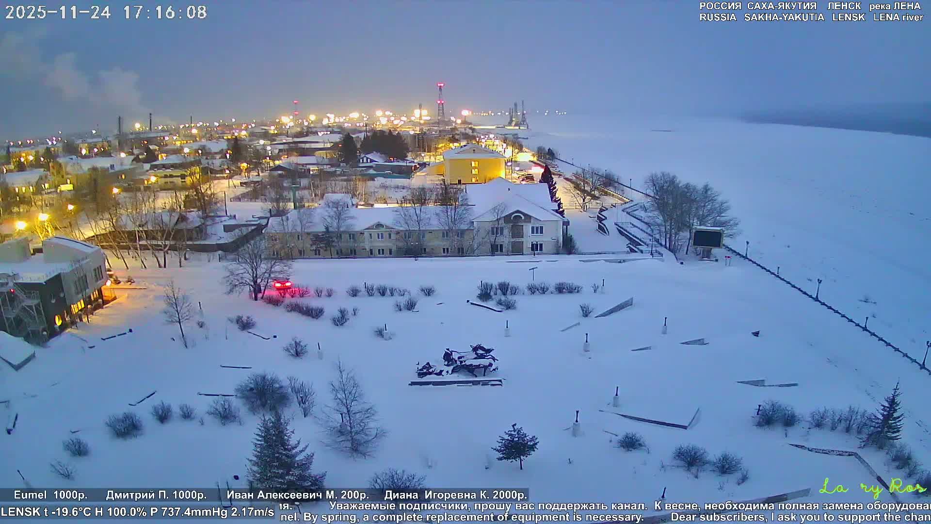 A snow-covered townscape with illuminated buildings and bare trees borders a frozen river under a dusky blue sky, revealing distant industrial lights in the cold winter evening.