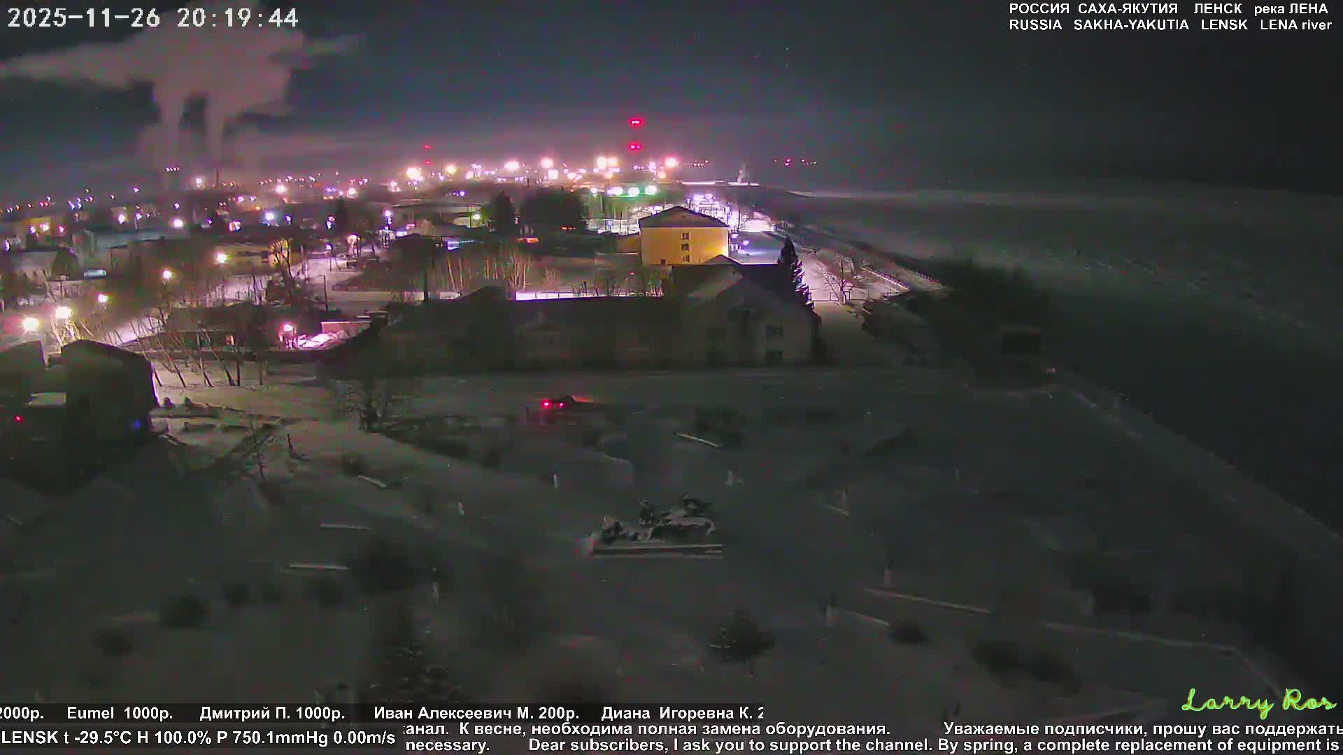 A snow-covered townscape with illuminated buildings and bare trees borders a frozen river under a dusky blue sky, revealing distant industrial lights in the cold winter evening.