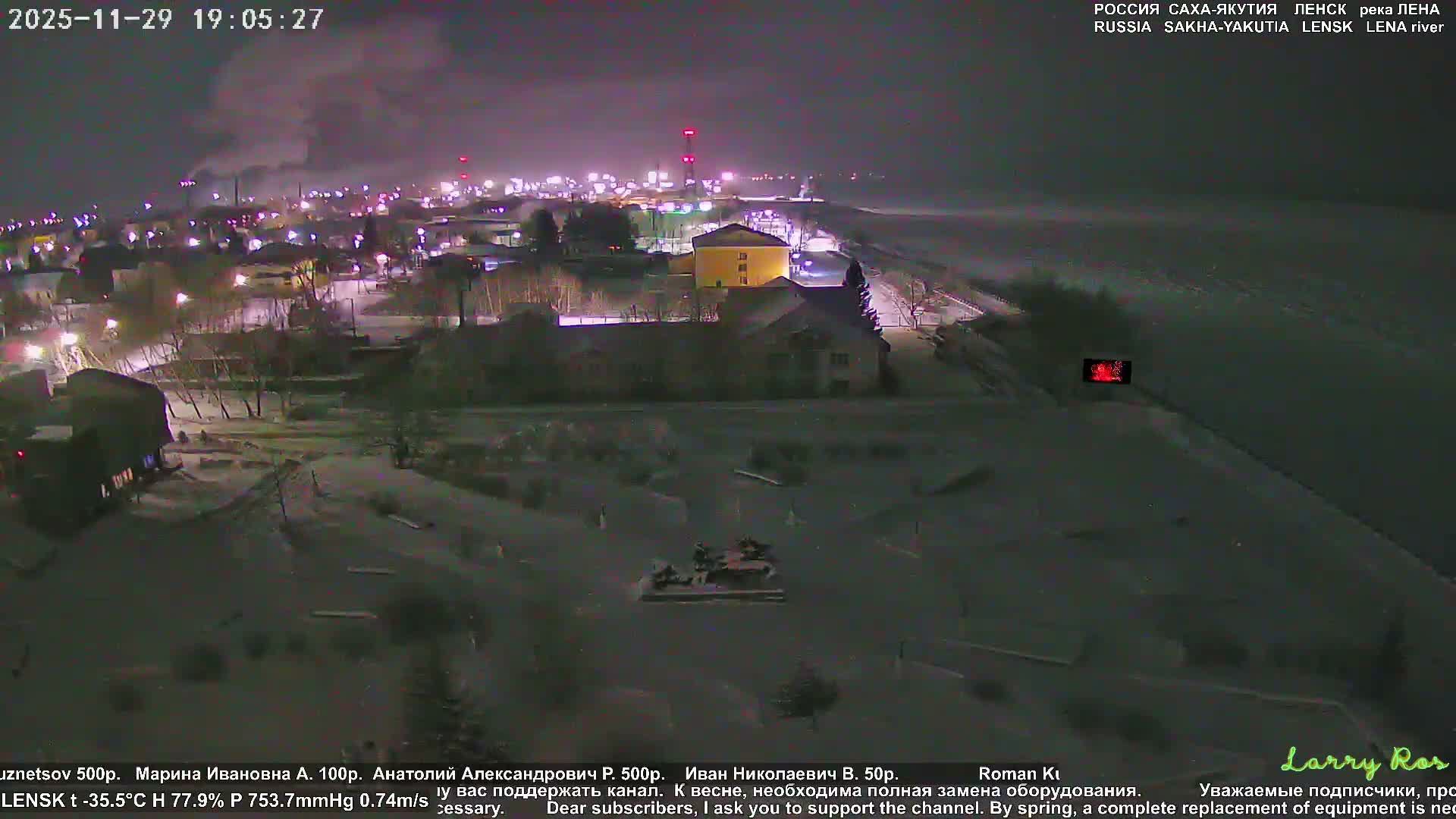 A frigid, hazy winter night reveals a snow-covered town illuminated by numerous lights, with buildings and bare trees bordering a wide, likely frozen river.