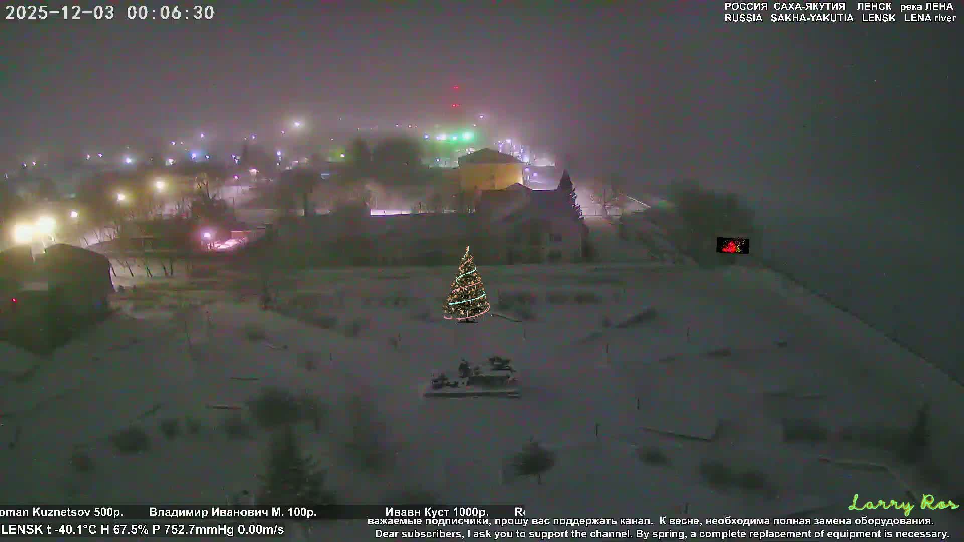 A brightly illuminated Christmas tree stands in a snow-covered outdoor landscape at night, with distant buildings and streetlights visible through what appears to be a very cold, hazy, or foggy atmosphere.