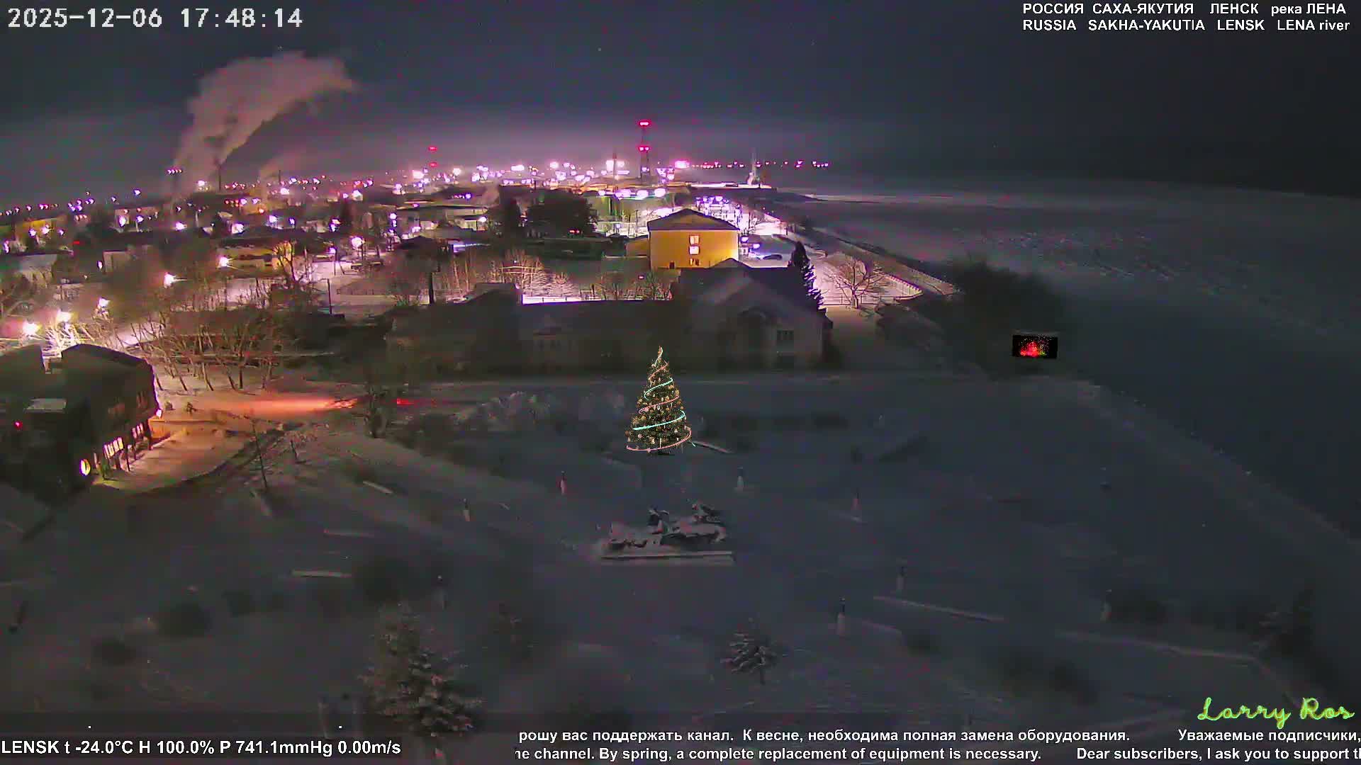 A snow-covered town shimmers with numerous street and building lights under a dark night sky, featuring a prominent decorated Christmas tree in the foreground and extending towards a vast, dark, frozen landscape to the right, indicating very cold winter conditions.