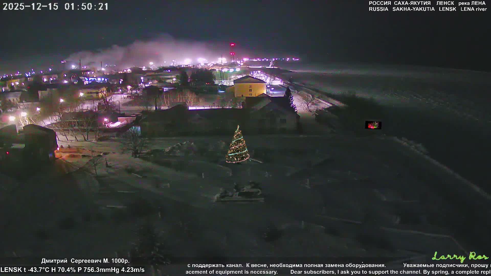 A snow-covered town shimmers with numerous street and building lights under a dark night sky, featuring a prominent decorated Christmas tree in the foreground and extending towards a vast, dark, frozen landscape to the right, indicating very cold winter conditions.
