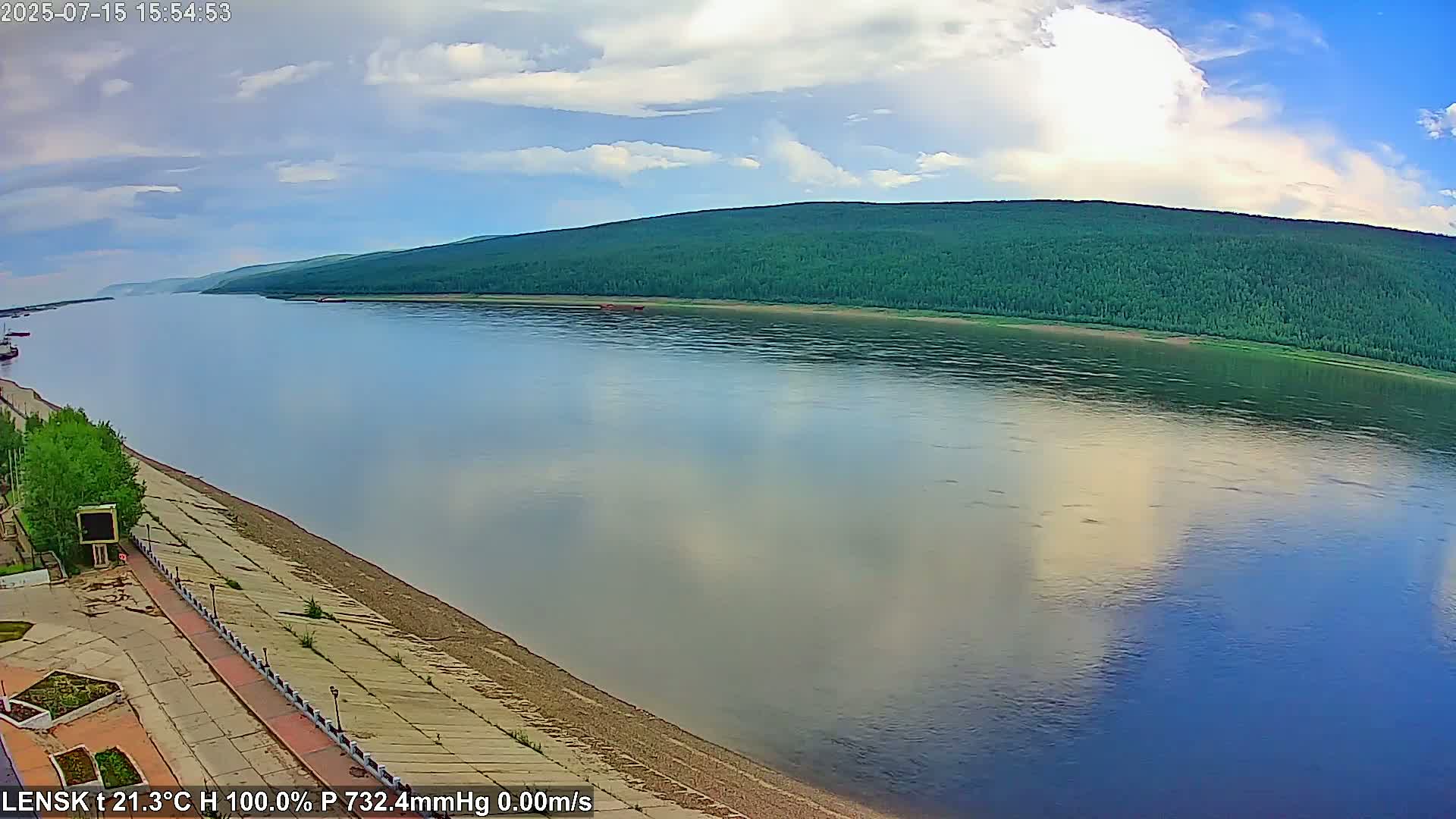 A wide, calm river reflects a partly cloudy sky under which a green, forested hill rises on the opposite bank, with a paved walkway along the river's edge in the foreground.