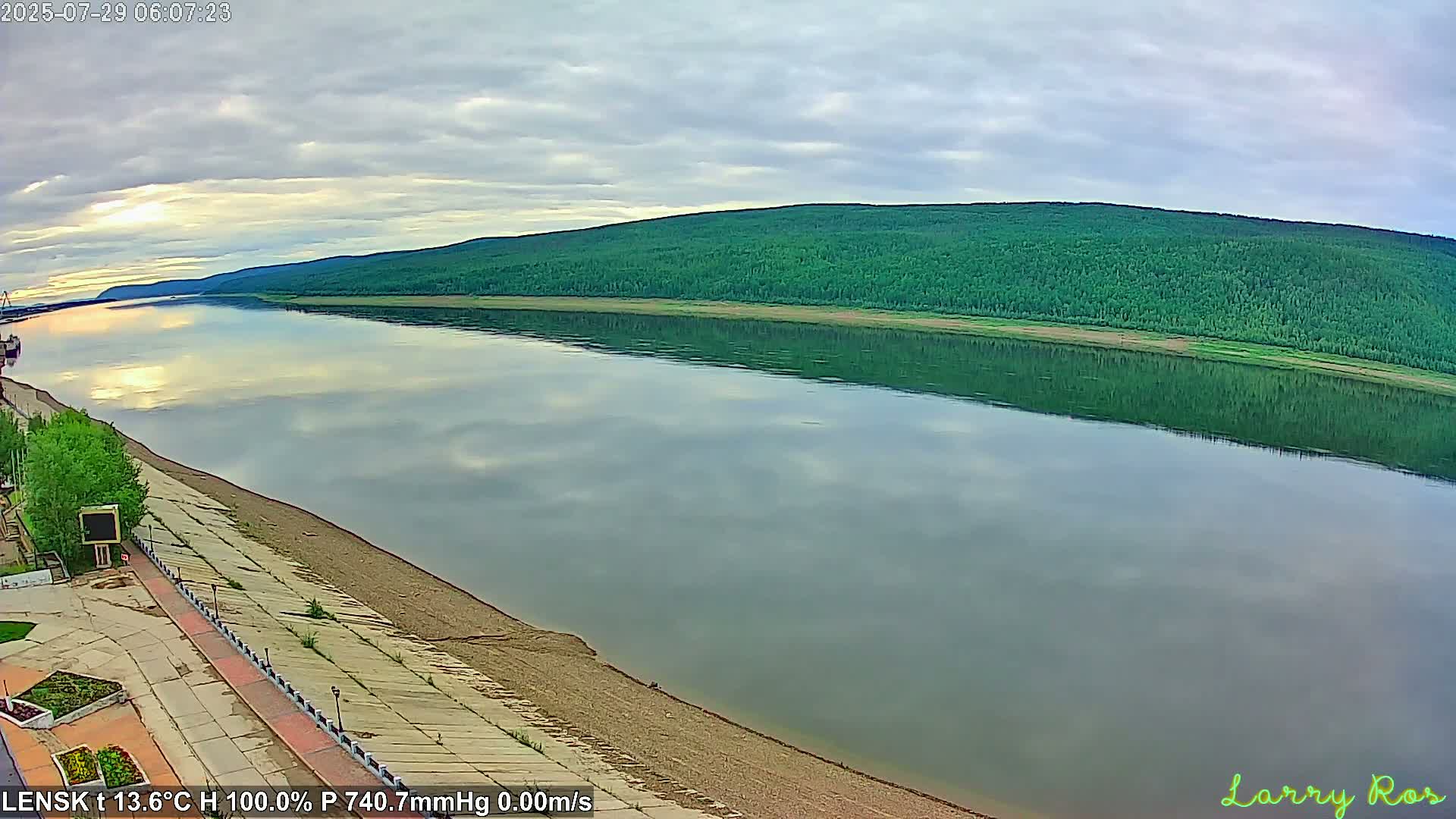 A calm river reflects a verdant hillside under a partly cloudy sky.
