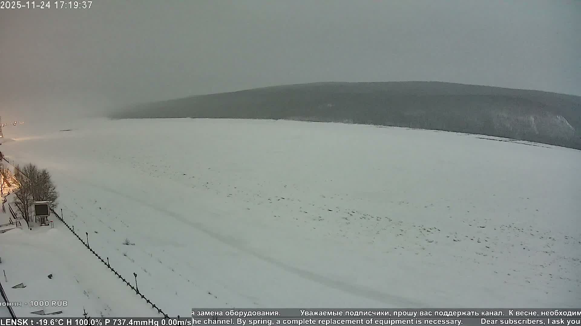 A vast, snow-covered plain, likely a frozen river or lake, stretches under a uniformly grey and overcast sky, with dark, hazy hills or a treeline in the distant background and sparse structures and bare trees visible on the left bank.