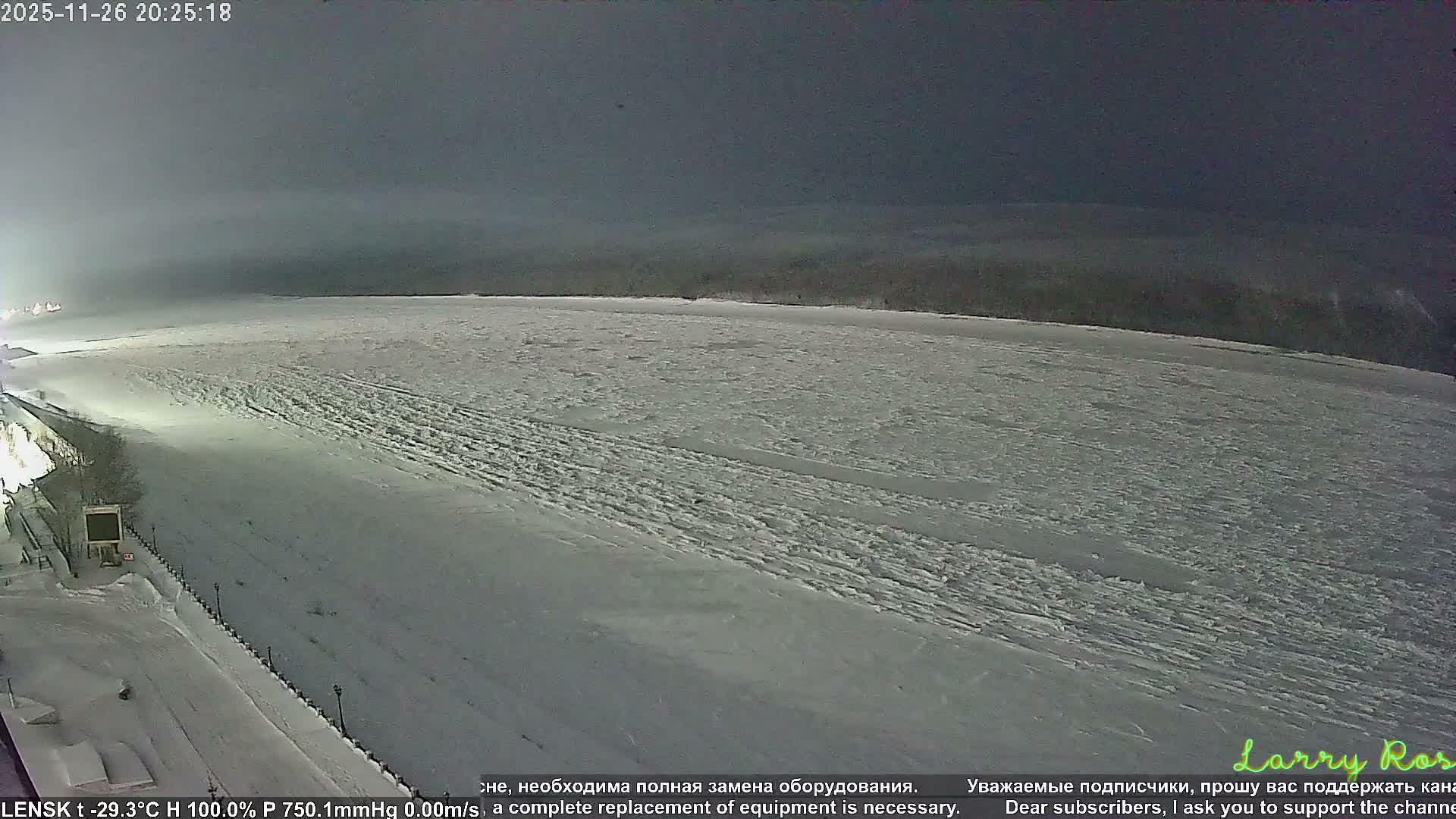 A vast, snow-covered plain, likely a frozen river or lake, stretches under a uniformly grey and overcast sky, with dark, hazy hills or a treeline in the distant background and sparse structures and bare trees visible on the left bank.