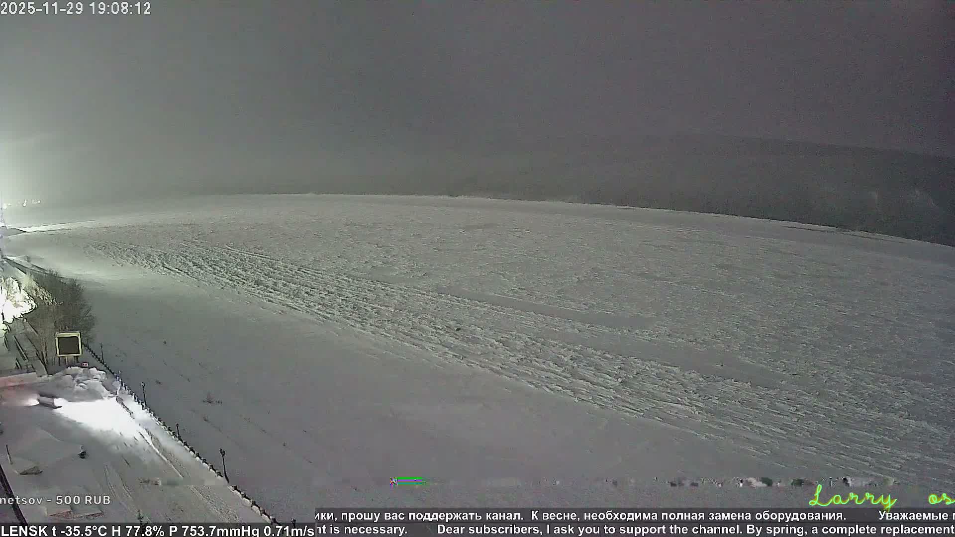 A vast, snow-covered landscape with visible tracks sprawls under a heavily overcast night sky, bordered on the left by a dimly lit road or embankment.
