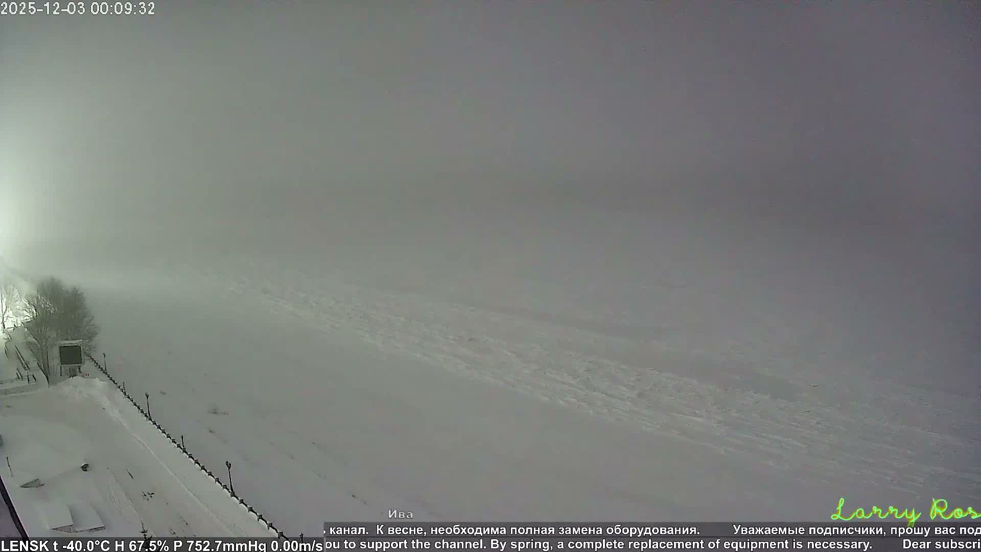 A snow-covered landscape at night is shrouded in thick fog or heavy overcast conditions, revealing bare trees and a line of dark posts along a slope on the left, with a bright, diffused light source in the distance.