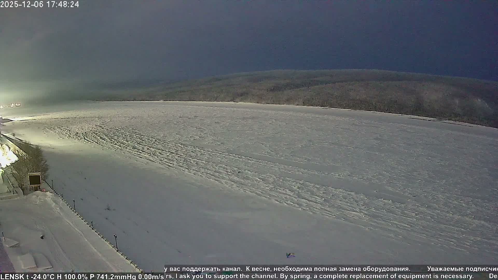 A wide, snow-covered winter landscape is seen at night, featuring a frozen expanse in the foreground, an illuminated embankment to the left, and a dark, forested horizon under a clear, dark sky.
