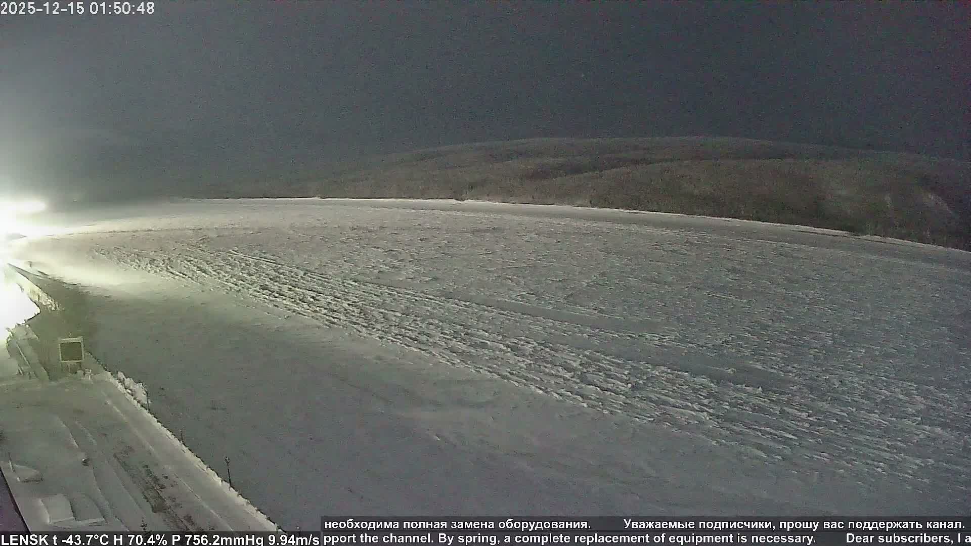 A wide, snow-covered winter landscape is seen at night, featuring a frozen expanse in the foreground, an illuminated embankment to the left, and a dark, forested horizon under a clear, dark sky.
