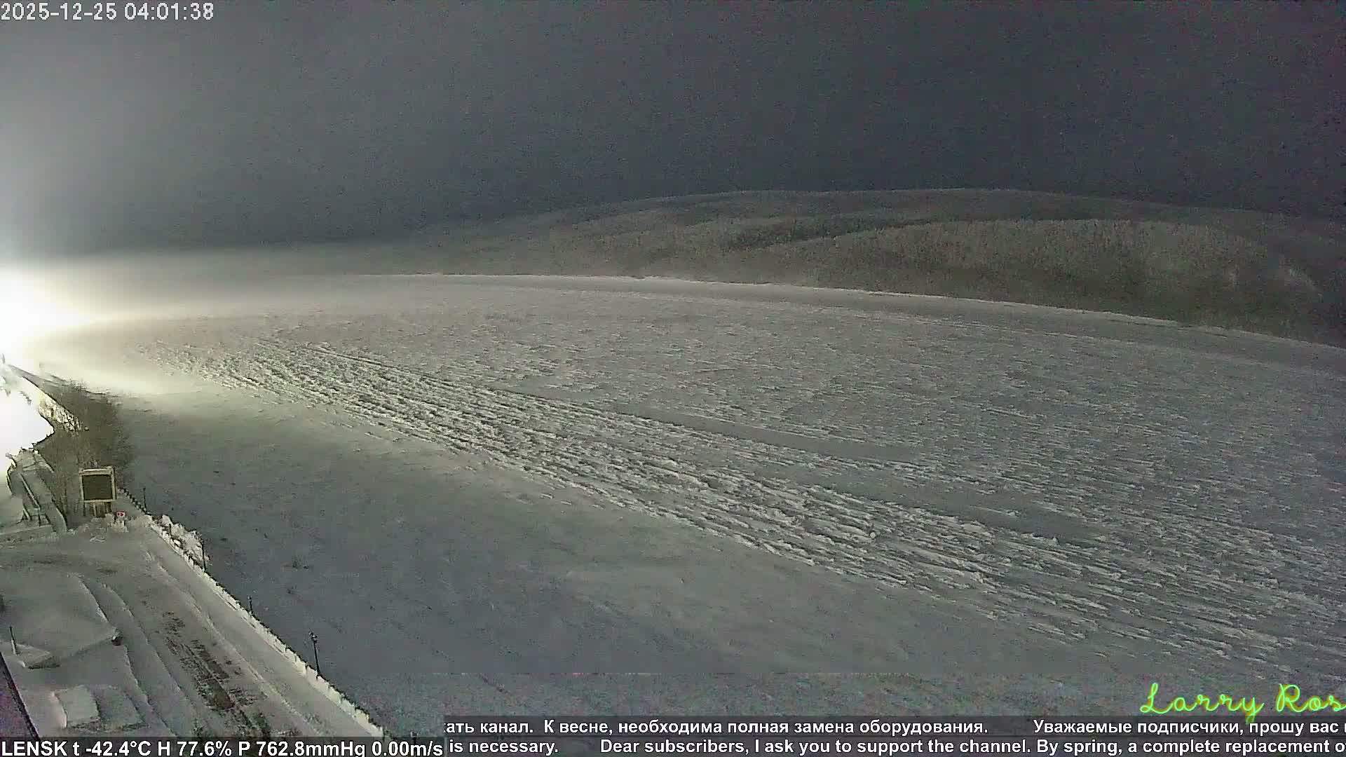A wide, snow-covered winter landscape is seen at night, featuring a frozen expanse in the foreground, an illuminated embankment to the left, and a dark, forested horizon under a clear, dark sky.