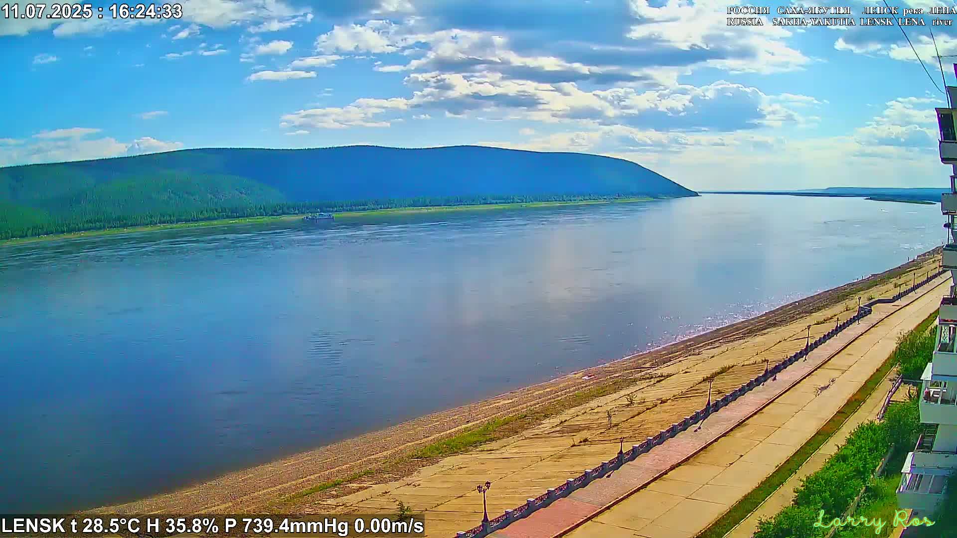 A wide, calm river flows past a sandy shoreline with a paved walkway and buildings under a partly cloudy sky.
