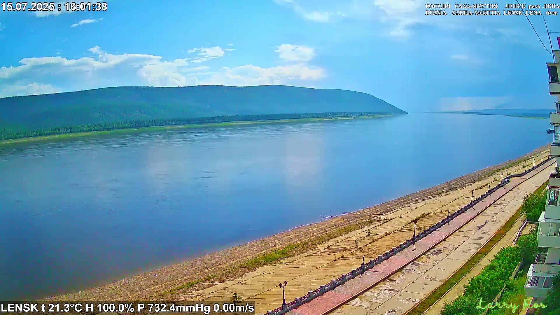 A wide, calm river flows past a paved waterfront path and a tree-lined hill under a mostly clear, bright sky.