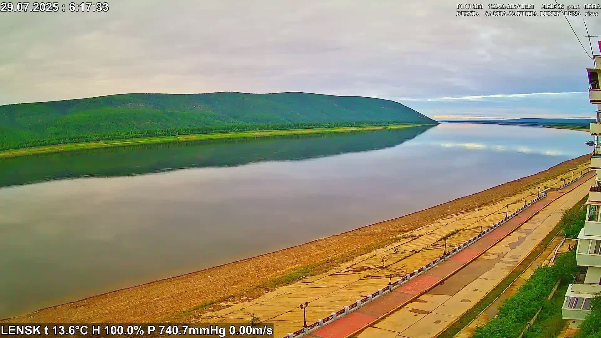 A wide, calm river reflects a green, tree-covered hill under a cloudy sky, alongside a paved waterfront walkway.
