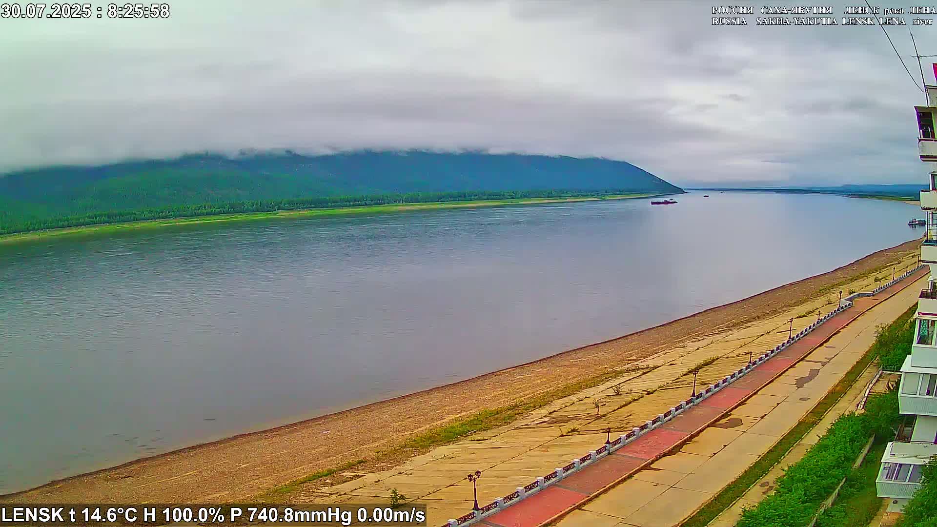 A wide, calm river reflects a green, tree-covered hill under a cloudy sky, alongside a paved waterfront walkway.