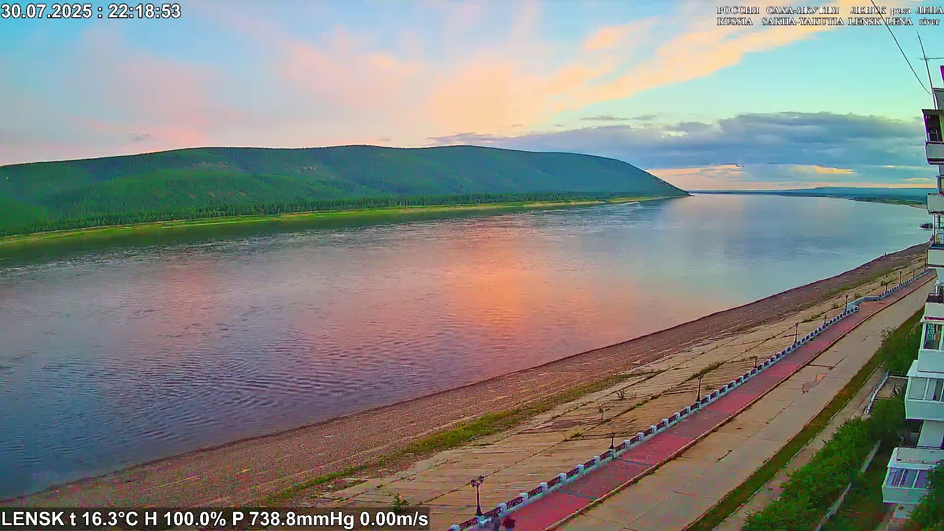A wide, calm river reflects a green, tree-covered hill under a cloudy sky, alongside a paved waterfront walkway.