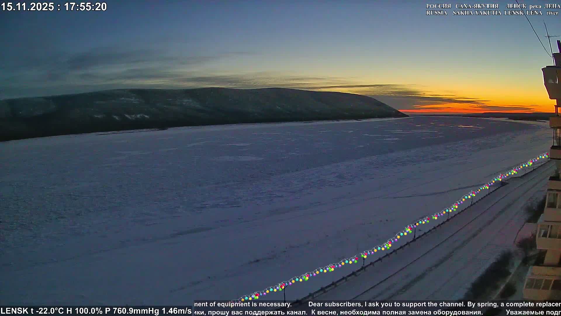 A wide, frozen river stretches towards distant snow-covered hills under a dramatic sunset sky with clear, very cold conditions, while colorful string lights line a visible bank in the foreground.