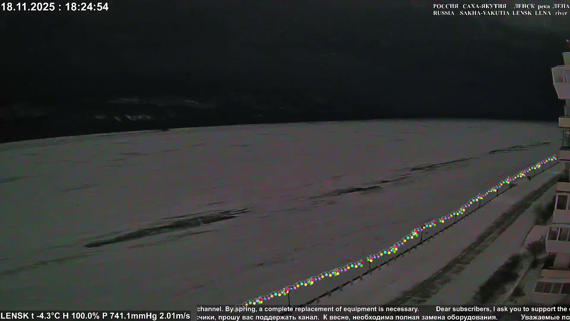 A wide, frozen river stretches towards distant snow-covered hills under a dramatic sunset sky with clear, very cold conditions, while colorful string lights line a visible bank in the foreground.