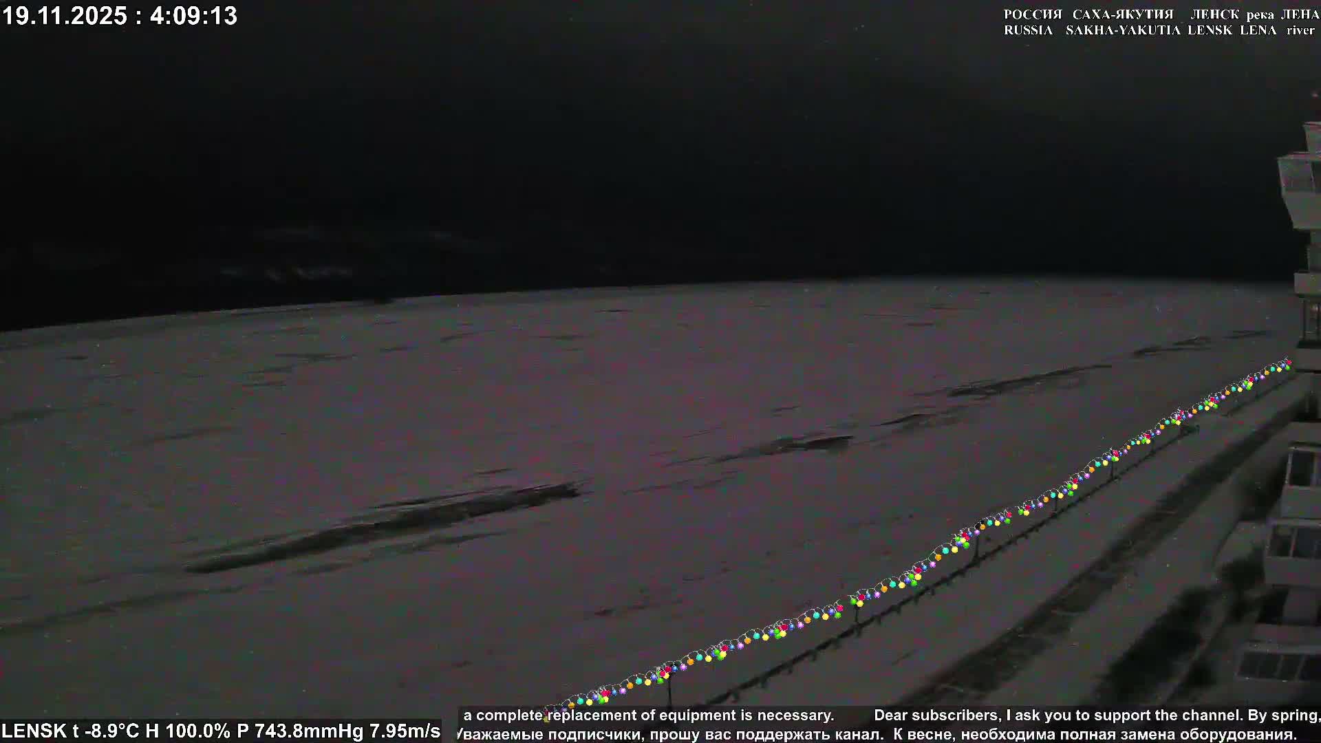 A wide, frozen river stretches towards distant snow-covered hills under a dramatic sunset sky with clear, very cold conditions, while colorful string lights line a visible bank in the foreground.