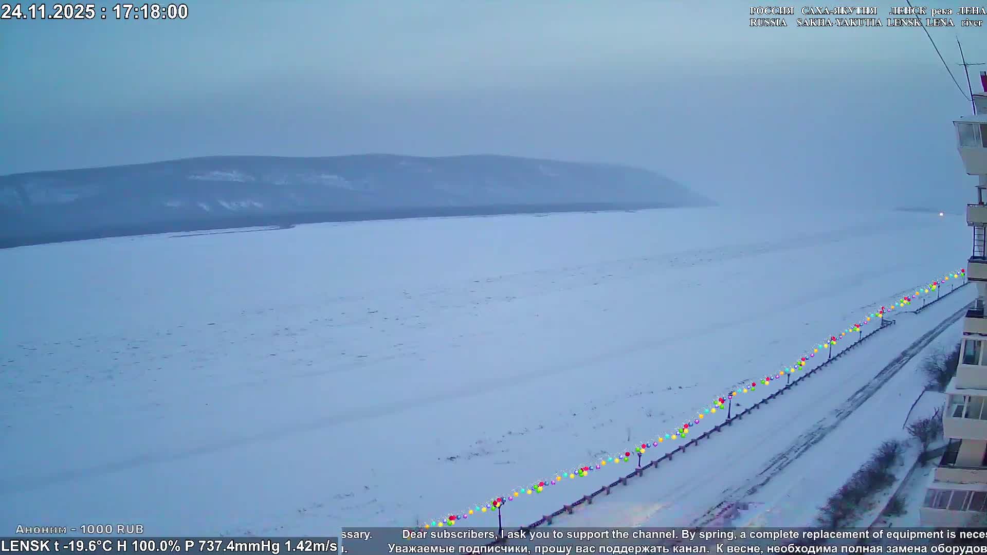 On a cloudy winter day, a vast frozen expanse covered in snow extends towards distant, muted hills under a pale sky, with a long line of colorful lights illuminating a snowy path beside a building in the foreground.