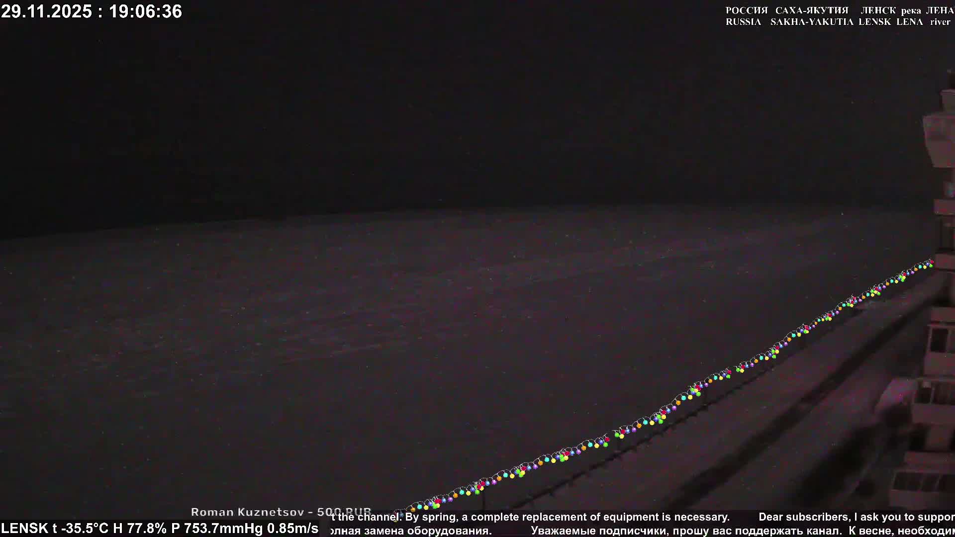A very dark outdoor scene at night with an extremely cold temperature of -35.5°C, featuring a long string of colorful lights extending diagonally across a dark foreground towards a faintly visible building on the right.