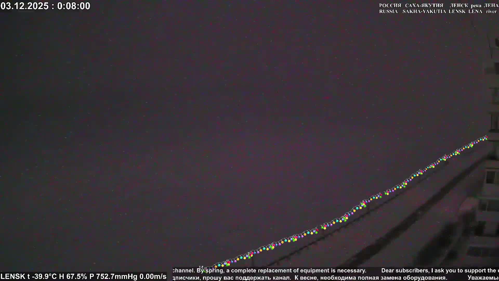 A very dark night scene features a long string of colorful lights spanning diagonally across the image, with a faint, dark building visible on the right, under a sky filled with numerous tiny, scattered bright dots, suggesting extremely cold, clear, or lightly precipitating conditions.