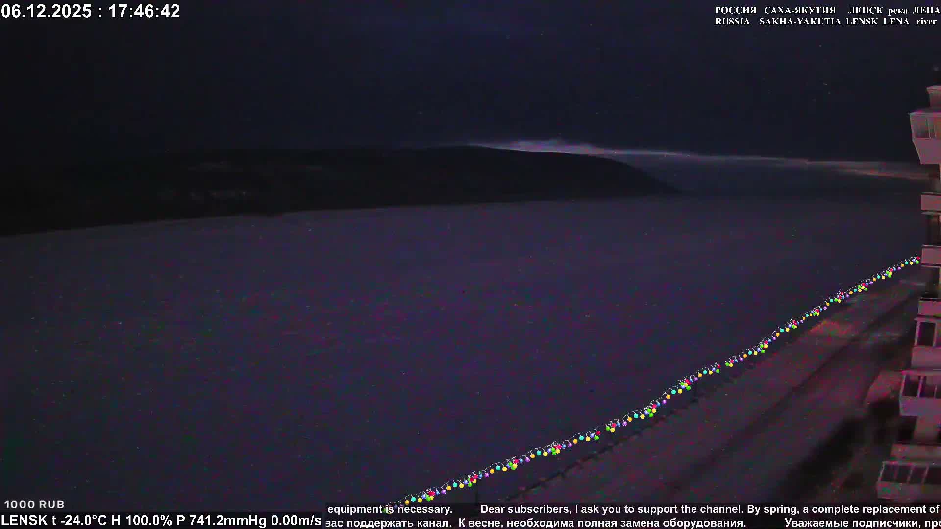 On a dark, cold winter night, a vast, frozen river or snow-covered landscape stretches under a deep blue sky with distant hills, while a string of colorful lights diagonally borders a building partially visible on the right.