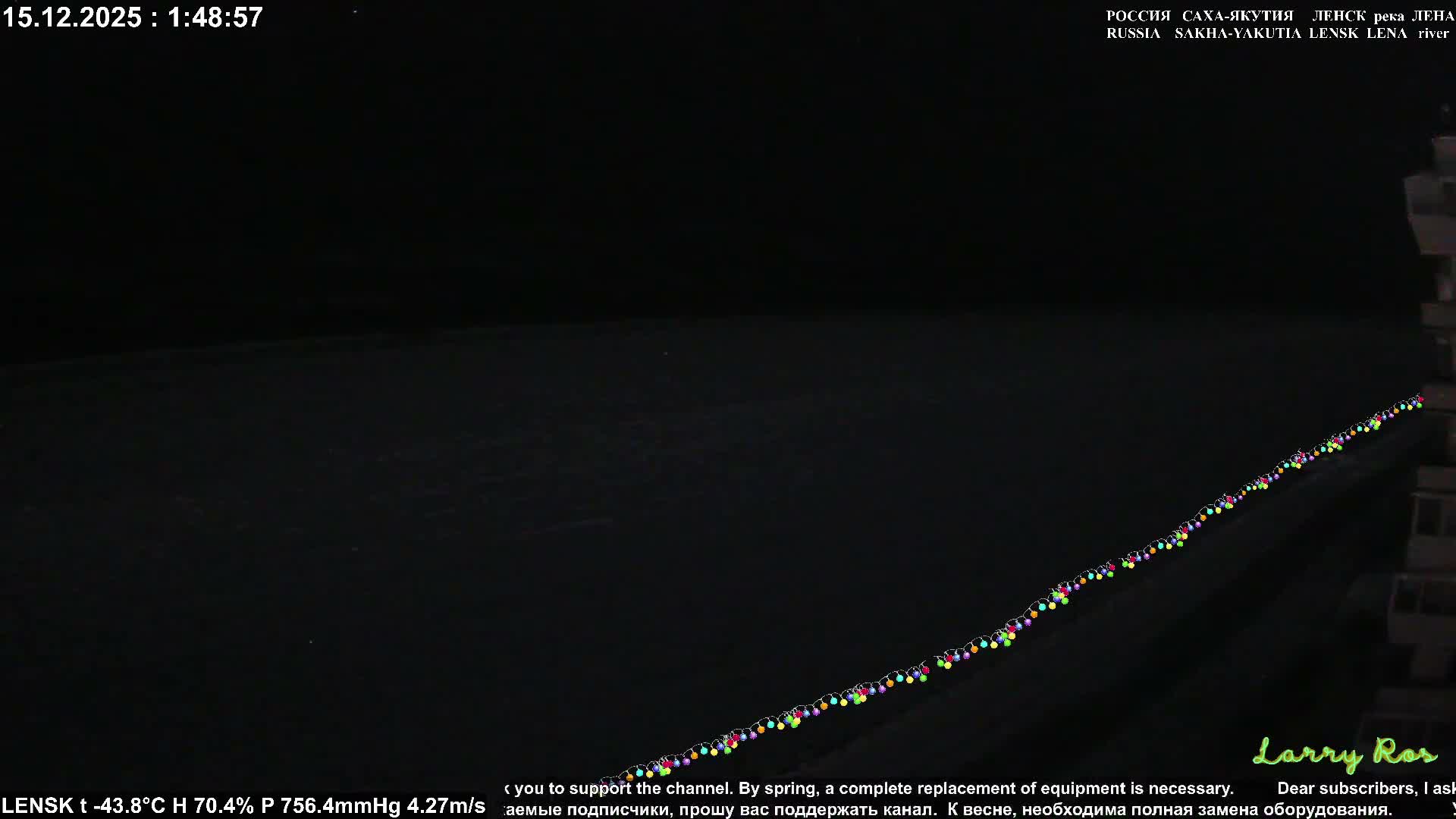 On a dark, cold winter night, a vast, frozen river or snow-covered landscape stretches under a deep blue sky with distant hills, while a string of colorful lights diagonally borders a building partially visible on the right.