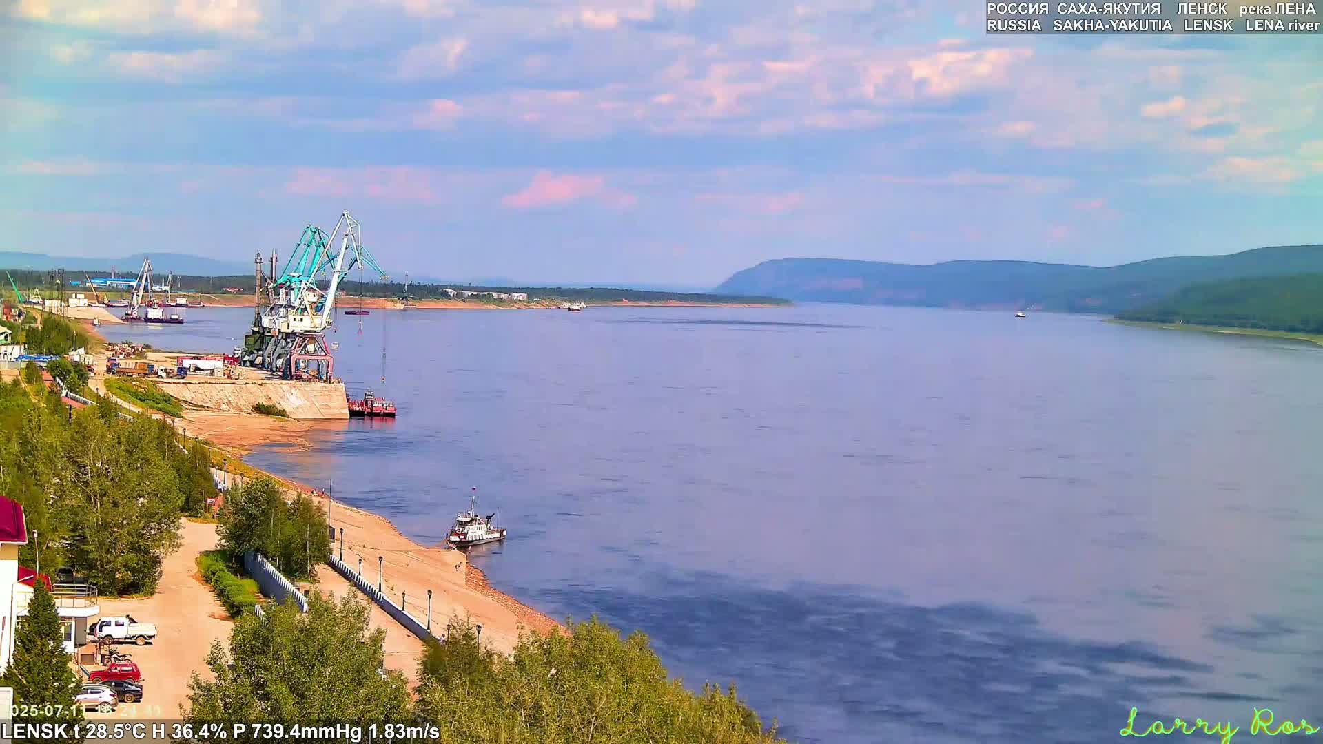 A wide, light-blue river flows past a sandy shore with a port featuring several cranes, a few small vessels, and a small town under a mostly clear sky.