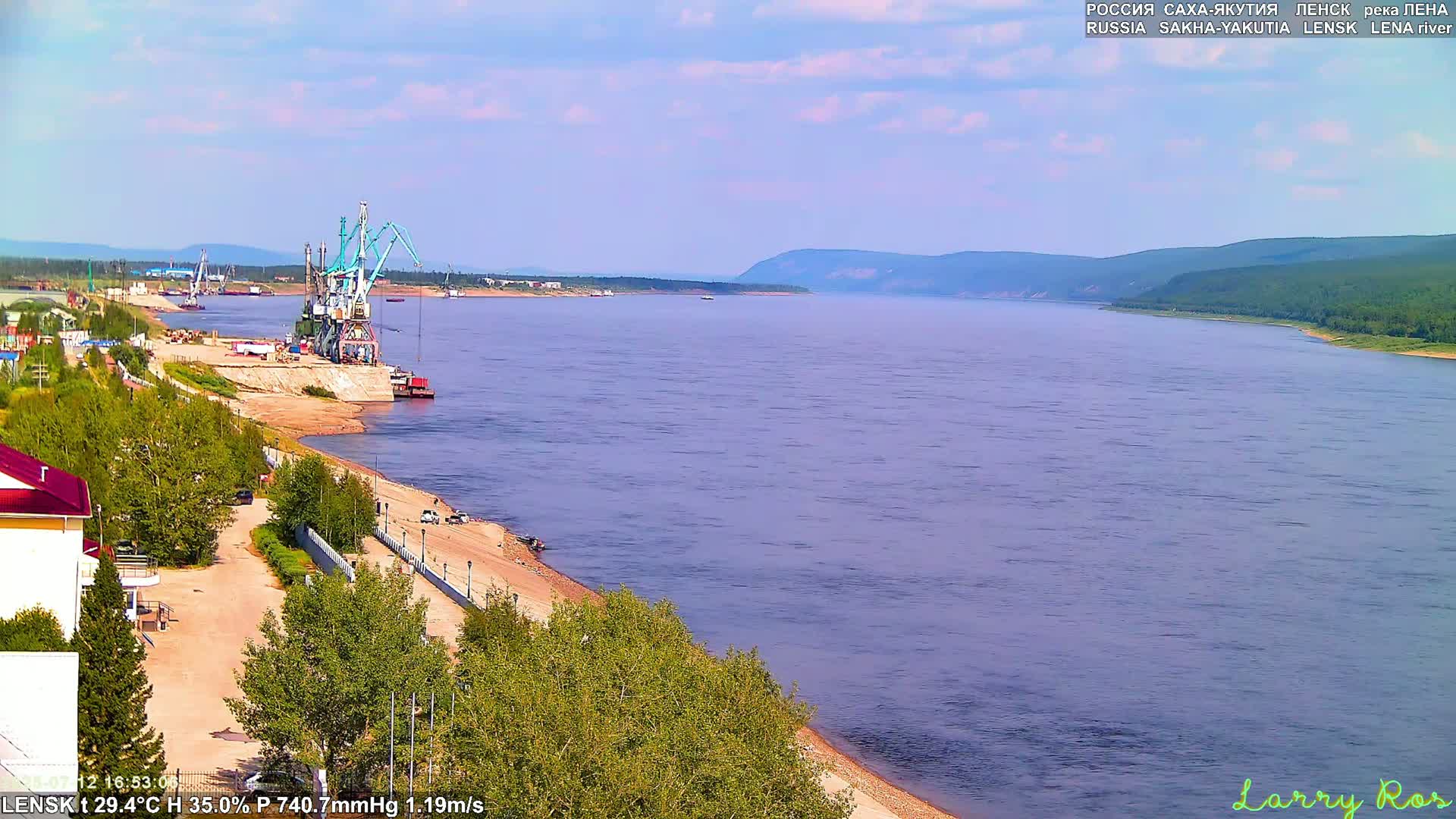 A wide, light-blue river flows past a sandy shore with a port featuring several cranes, a few small vessels, and a small town under a mostly clear sky.