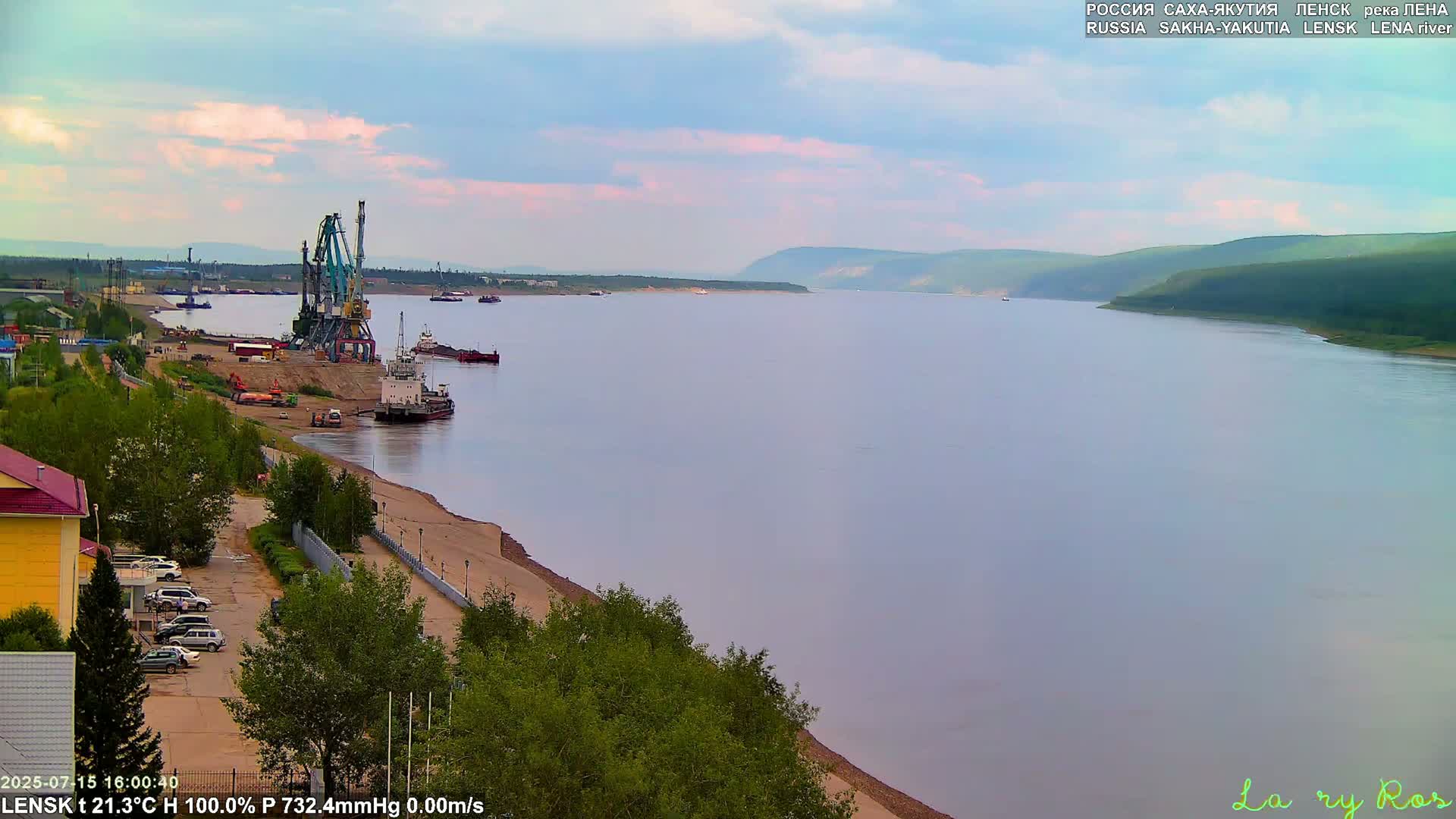 A wide, calm river flows past a shoreline with a port, featuring cranes and several vessels, under a partly cloudy sky.
