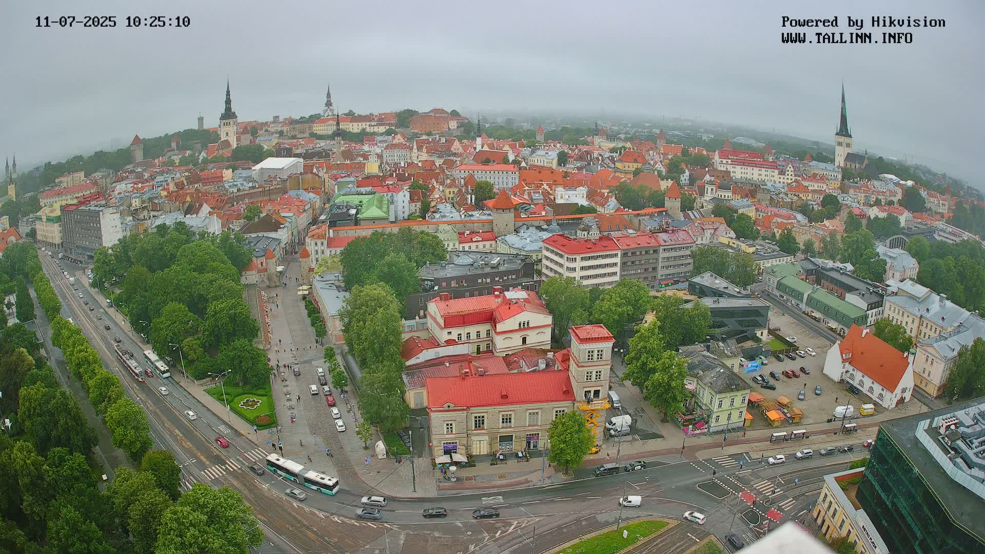 An aerial view of a city on an overcast day shows a large expanse of low buildings with red roofs, several streets with traffic, and green trees.